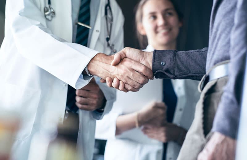 person wearing lab coat shakes another person hand, woman in background