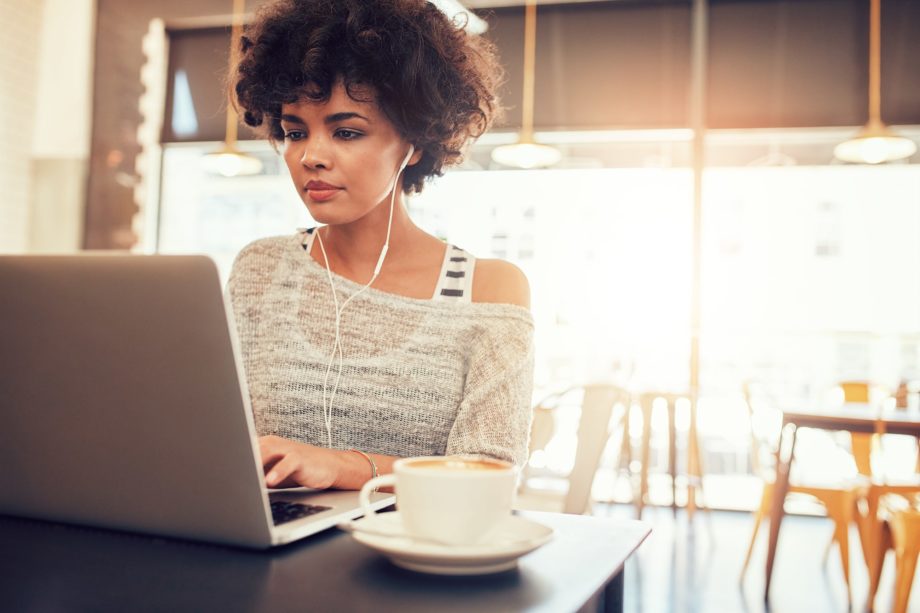 Portrait of beautiful young woman with earphones working on laptop while sitting at a coffee shop. African female at a cafe surfing internet on laptop.