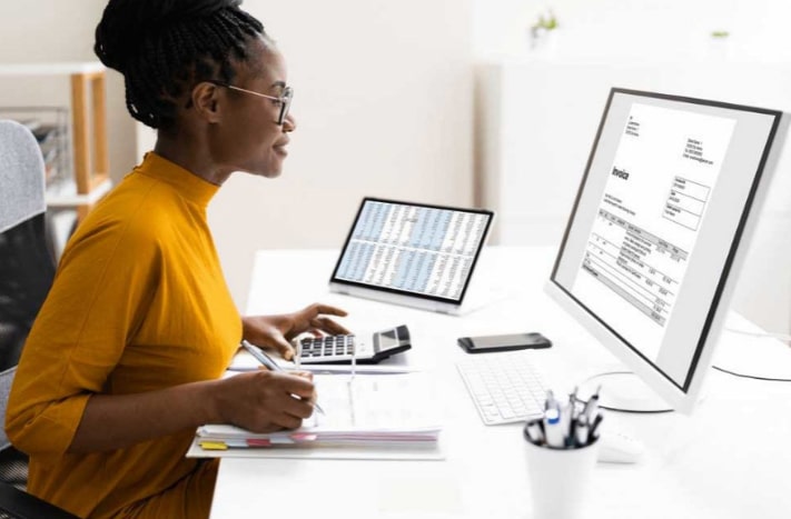 Woman sitting at a desk looking at a medical invoice on a computer monitor.