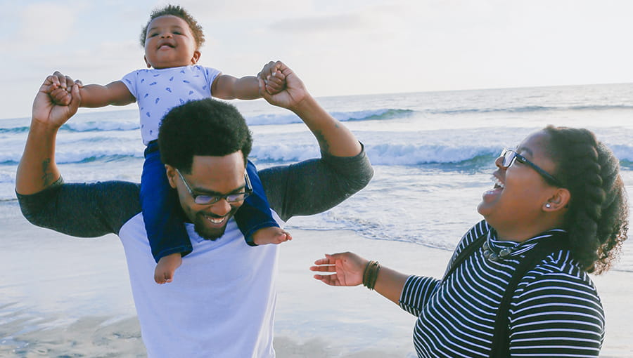 A couple on the beach with the father carrying the baby on his shoulders while mother laughs